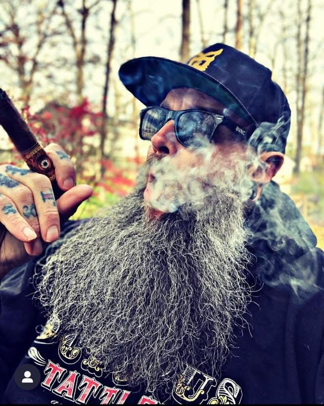 Man with a long beard and sunglasses smoking a pipe in a forest setting